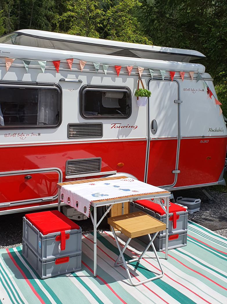 Vor einem Camper stehen ein Tisch sowie Sitzgelegenheiten aus Seat Boxen.